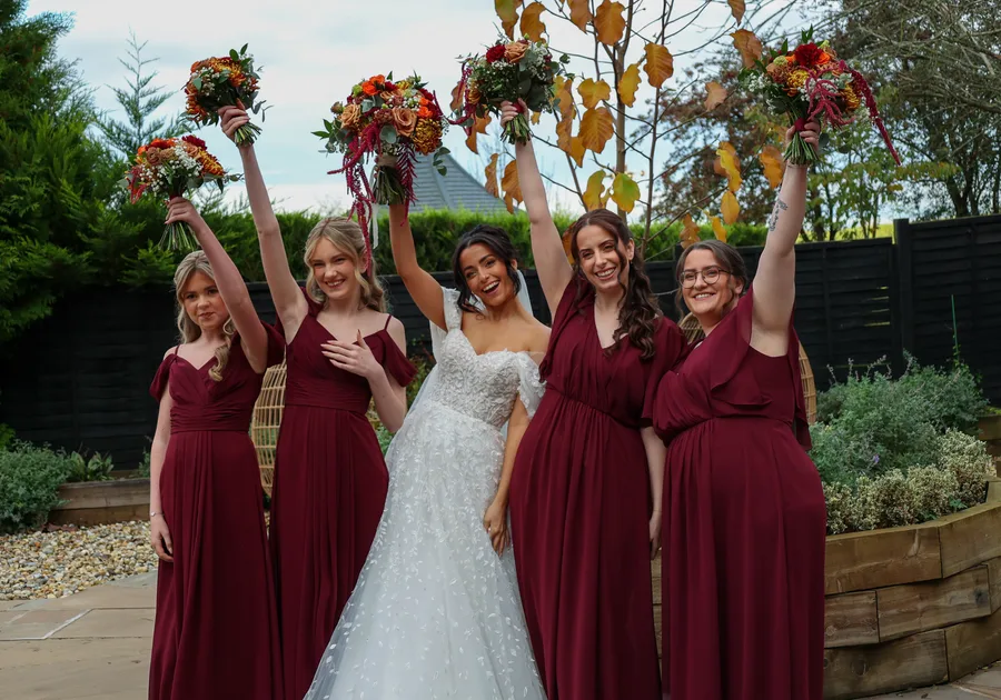 Bridesmaids in burgundy raising their autumn bouquets with the bride