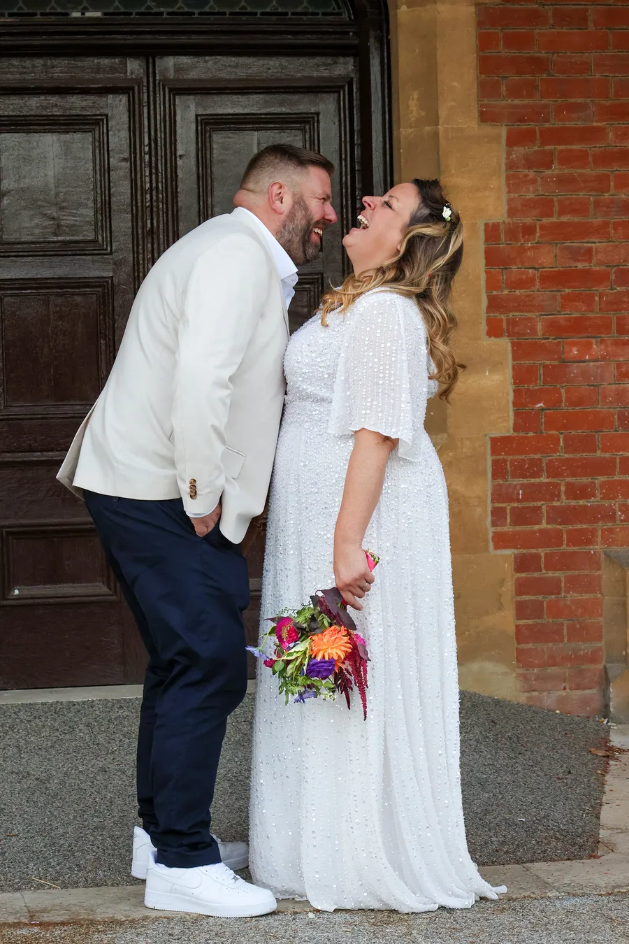 Bride and groom leaning together laughing outside a red-brick venue, the bride holding a bright autumn bouquet