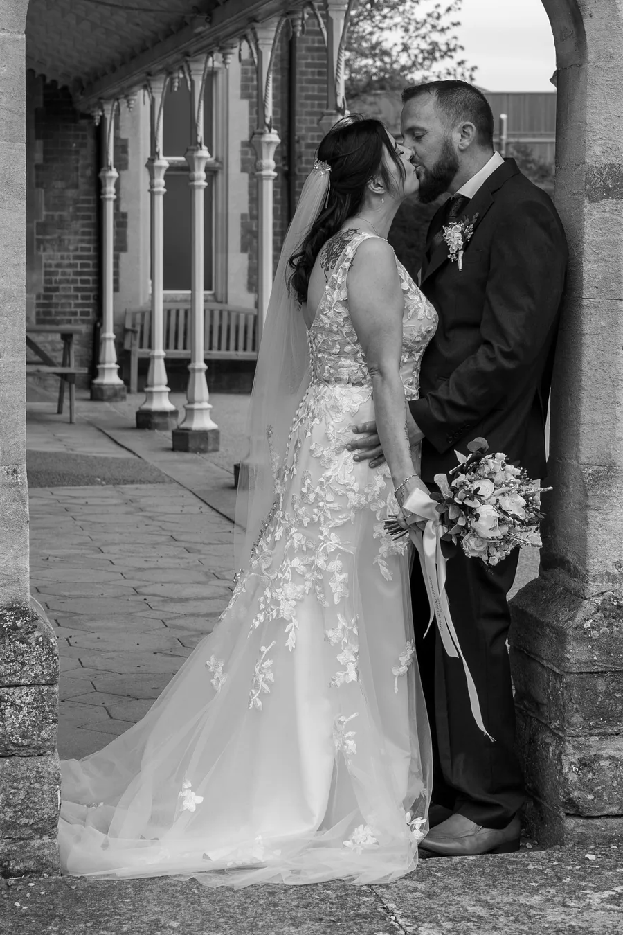 Bride and groom kissing under a brick colonnade, in black and white