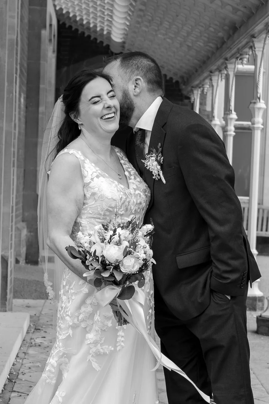 Groom kissing the bride's cheek as she laughs, in black and white