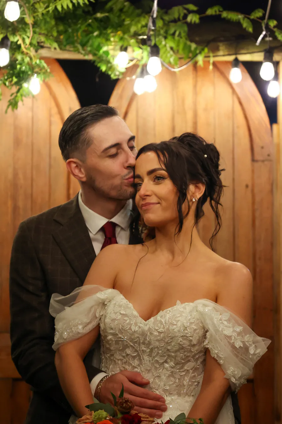Groom kissing the bride's cheek under festoon lights