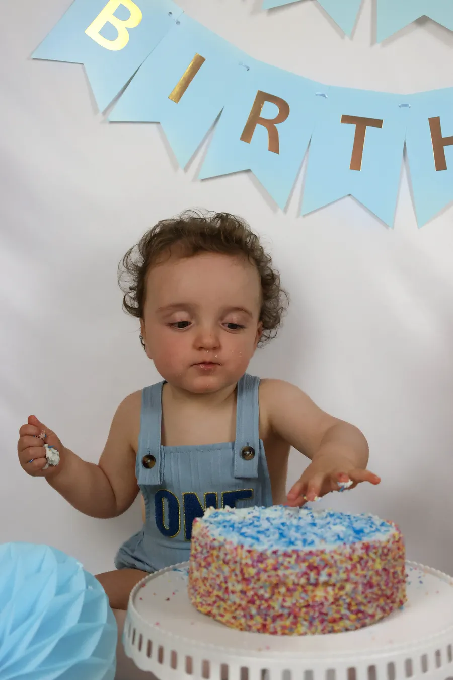 One-year-old in dungarees reaching for his sprinkle-covered birthday cake