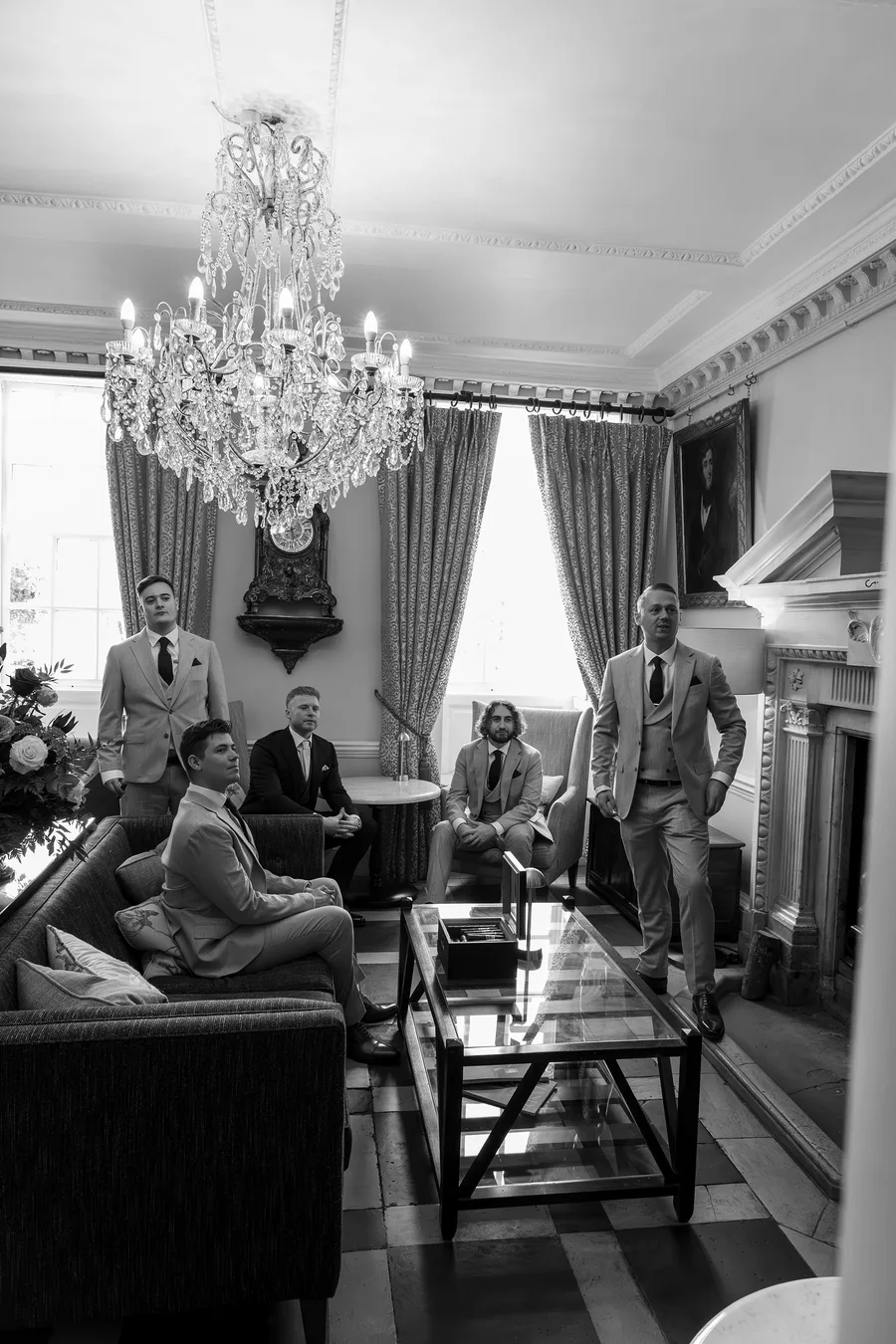 Groomsmen waiting in the drawing room beneath a chandelier, in black and white