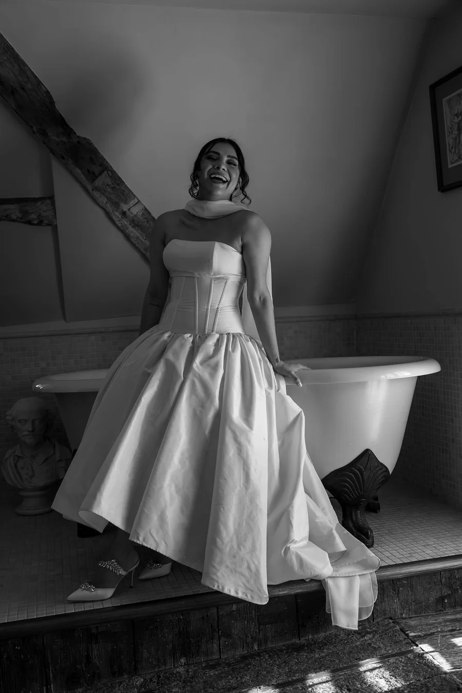 Bride sitting on a roll-top bath in her gown, in black and white