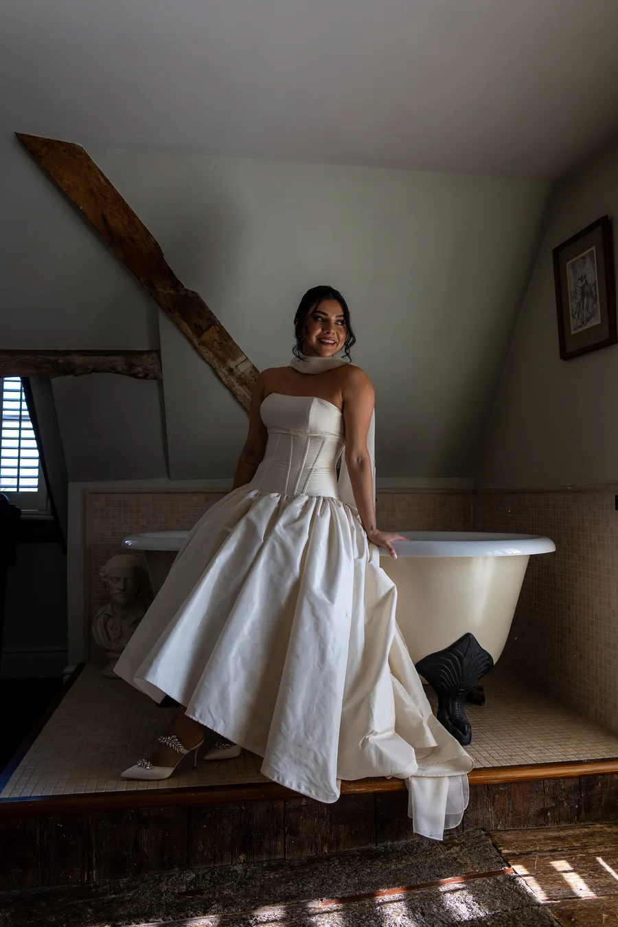 Bride in her gown beside a roll-top bath under the eaves