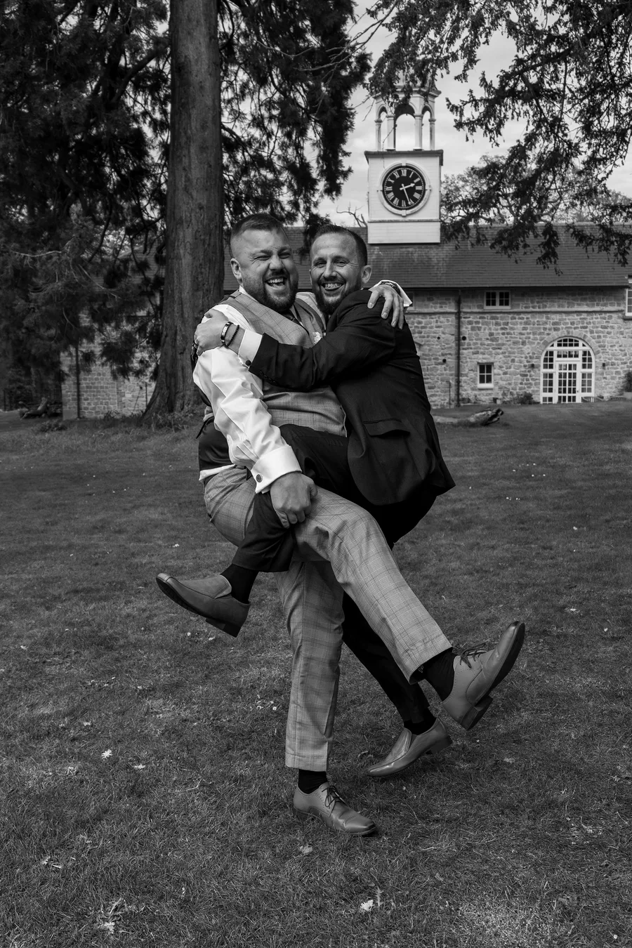 A groom lifted up by his best man on the lawn, in black and white