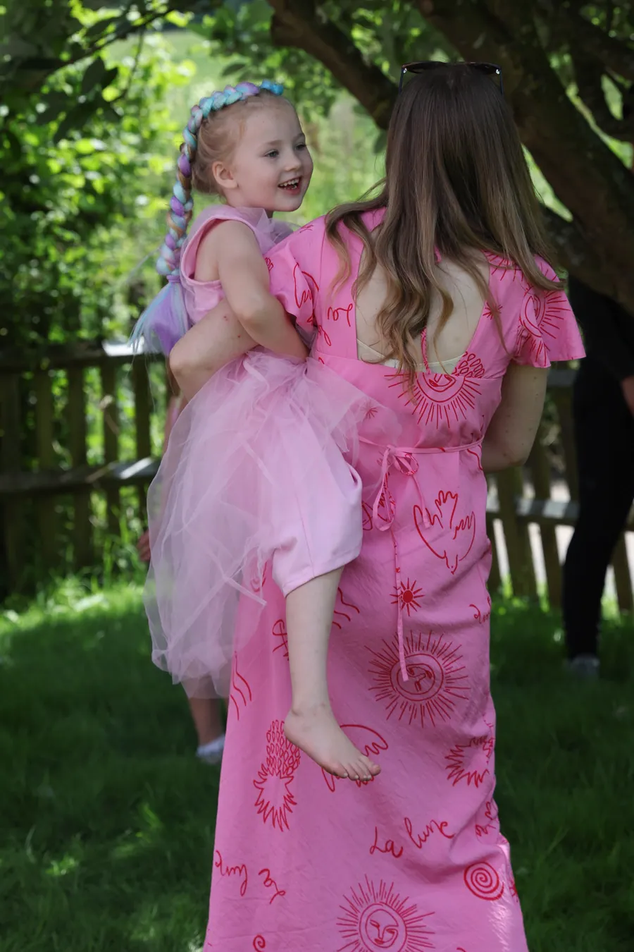 A woman carrying a little girl in a pink party dress through the garden